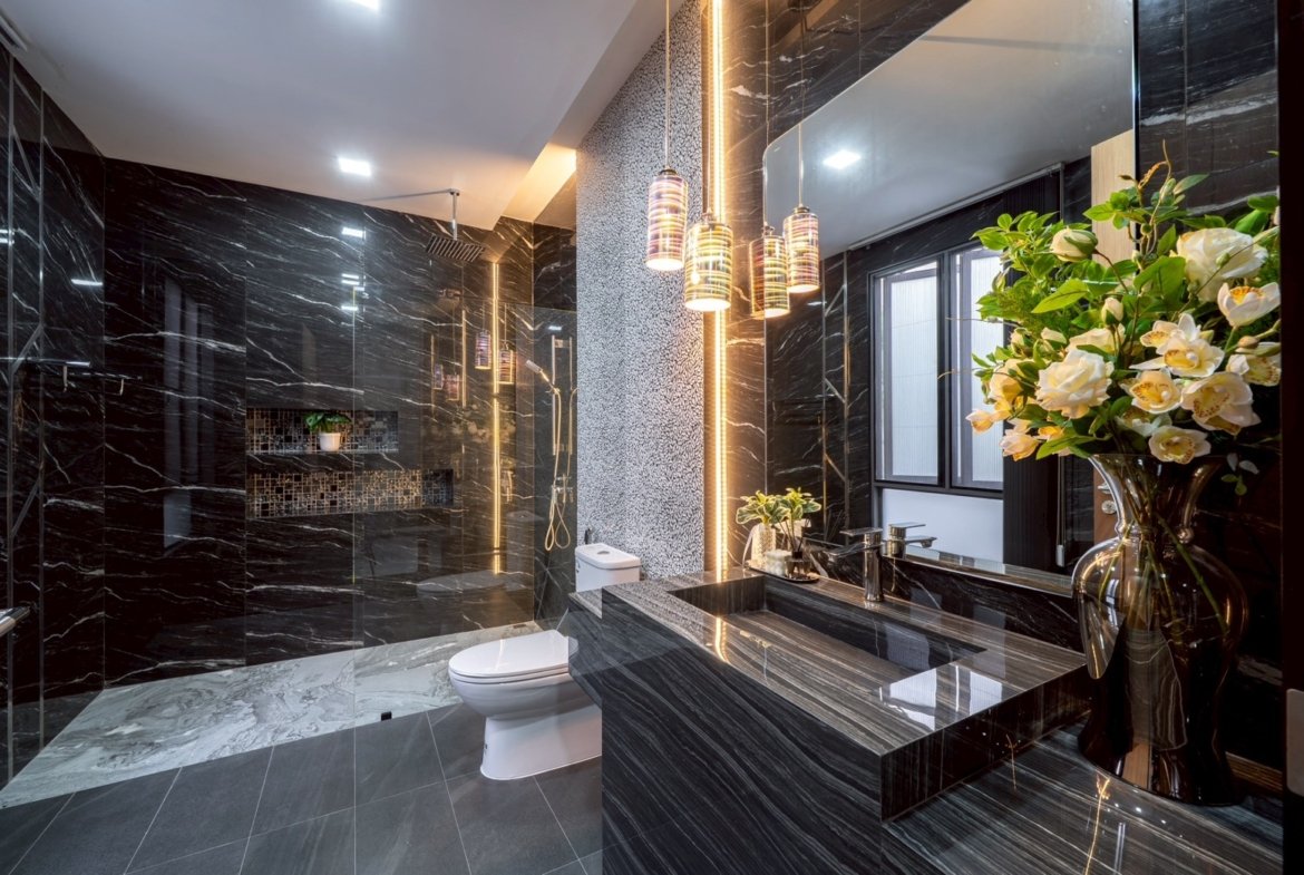 Luxurious black marble bathroom with glass shower, wall-to-wall mirror, and a dark marble vanity topped with a vase of white flowers.