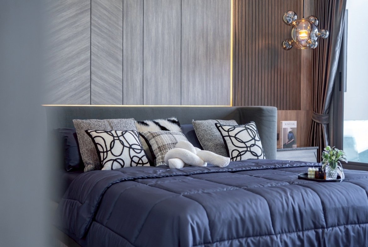 Luxurious modern bedroom with a dark blue comforter and a row of patterned throw pillows on a padded headboard, light gray wall panels, and a glass bubble chandelier near a window.