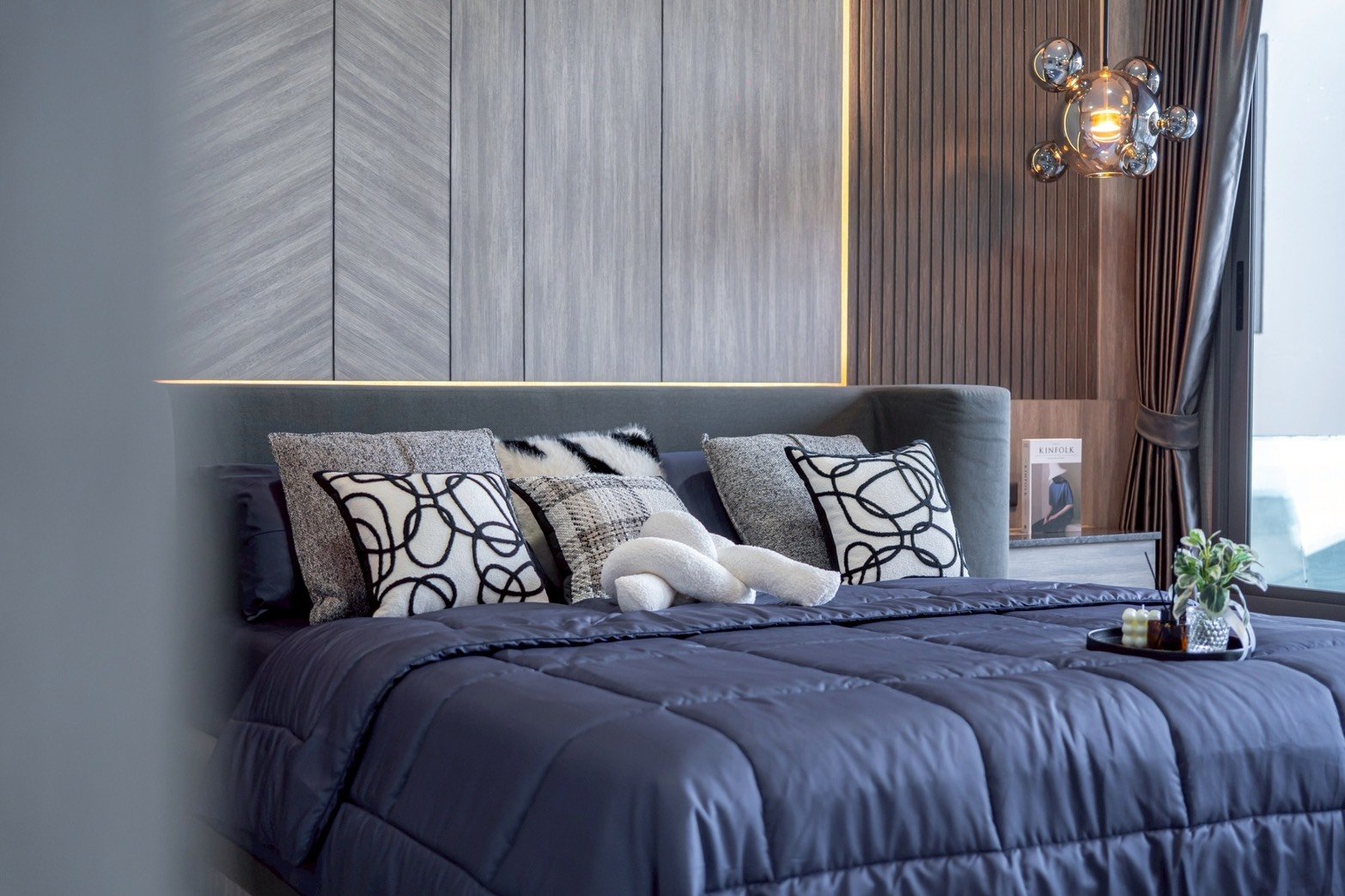 M_Mountain_Dlh518 32 Luxurious modern bedroom with a dark blue comforter and a row of patterned throw pillows on a padded headboard, light gray wall panels, and a glass bubble chandelier near a window.