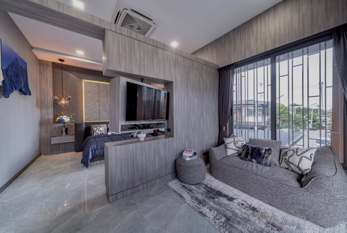 Modern hotel suite with a grey wood-paneled living area, wall-mounted TV, and an open bedroom separated by a partial wall and wardrobe-looking structure, plus large balcony doors.