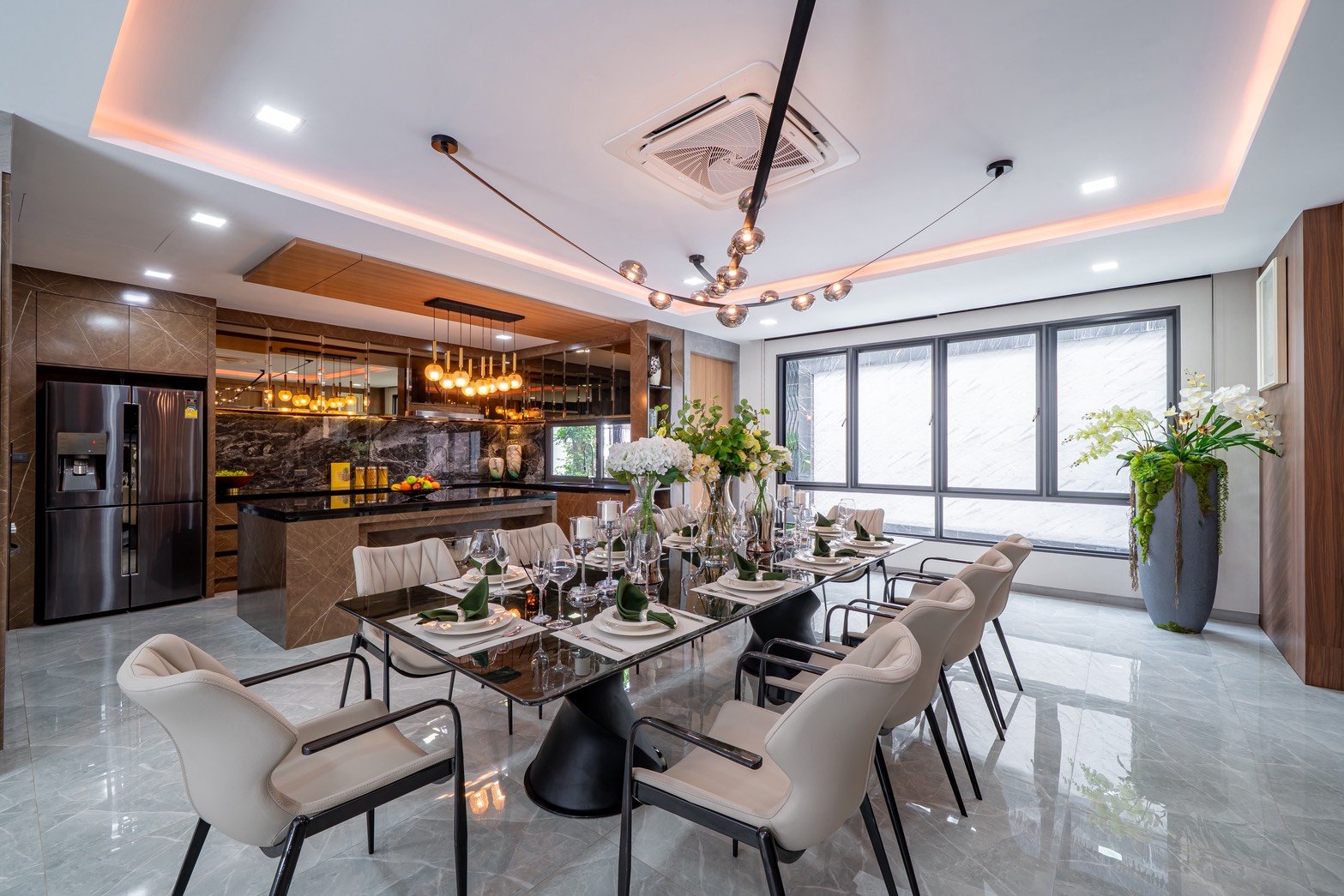 M_Mountain_Dlh518 4 Modern dining room with a long glass table set for a formal meal, cream upholstered chairs, and floral centerpieces under warm ambient lighting.