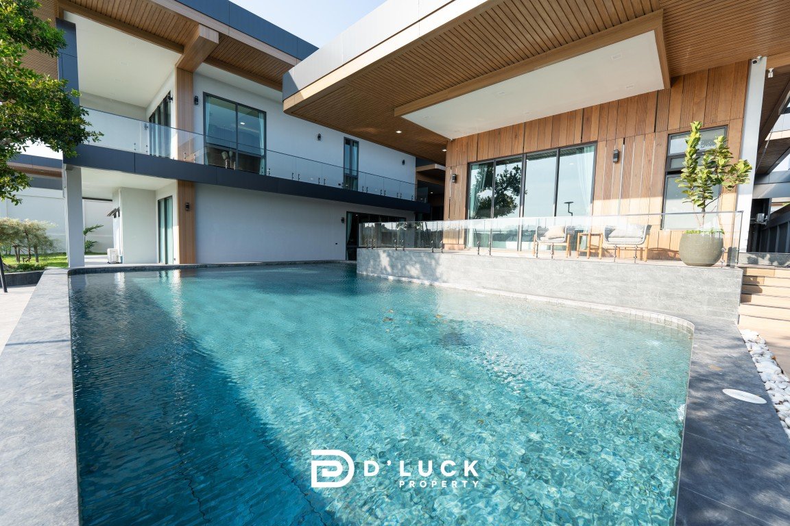 Modern two-story home with an expansive turquoise pool, glass railings, and wooden exterior accents; outdoor seating area visible beyond the pool.