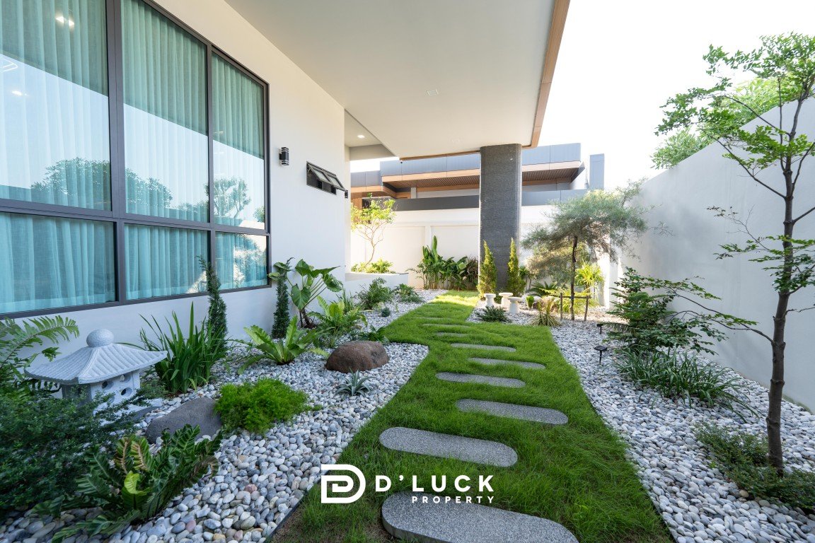 Modern courtyard with a stepping-stone path on a green lawn, surrounded by white gravel and tropical plants beside a glass-walled building.