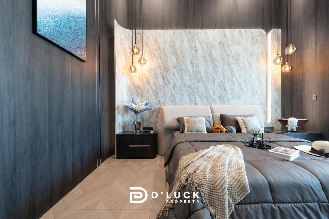 Modern bedroom with a gray upholstered bed, marble feature wall, warm pendant lights, and dark nightstands; D’Luck Property logo at the foot of the bed.