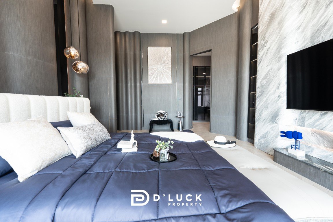 Luxurious modern bedroom with a navy quilted bed, white headboard, gray paneled walls, and a mounted flat-screen TV across a marble-accent wall. Watermark reads D’LUCK PROPERTY.