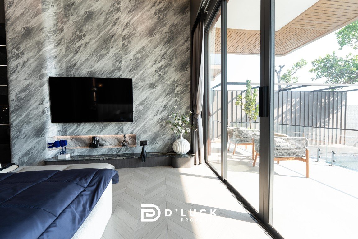 Modern bedroom with a marble accent wall, wall-mounted TV, and a blue quilted bedspread; glass doors open to a sunny balcony.