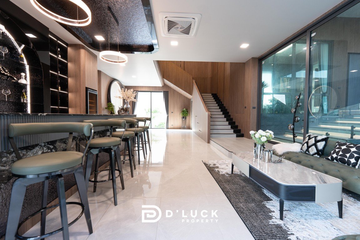 Luxurious open-concept living area with a long bar, green upholstered stools, and marble floors; stairs and glass wall visible beyond.