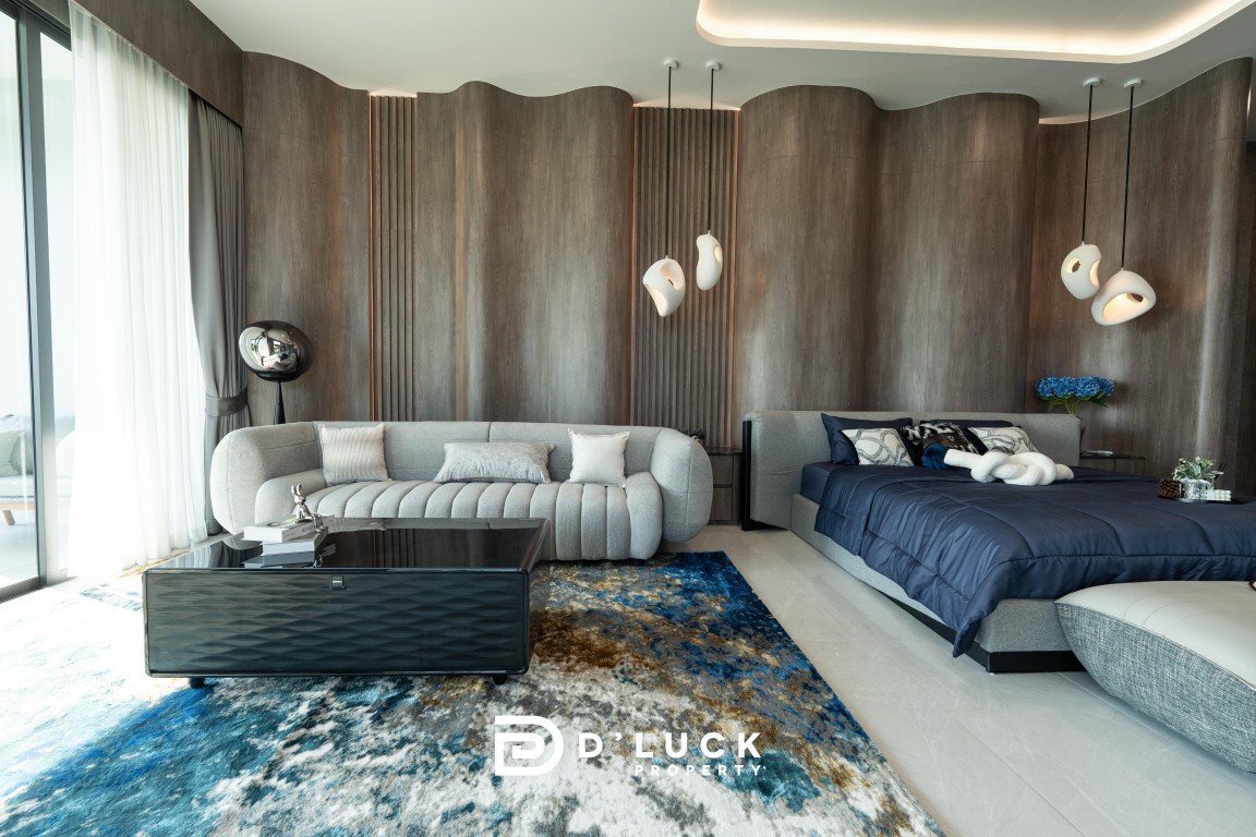 Luxurious hotel suite with a curved gray sofa, navy bed, glass coffee table, and a blue-toned abstract rug against a wood-paneled wall with pendant lights.