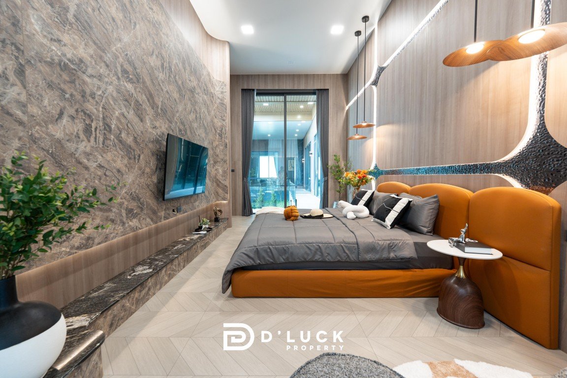 Contemporary hotel bedroom with a gray platform bed, orange headboard, and a marble accent wall; a wall-mounted TV and long marble ledge run along the left, with a balcony door at the far end and plants for decor.