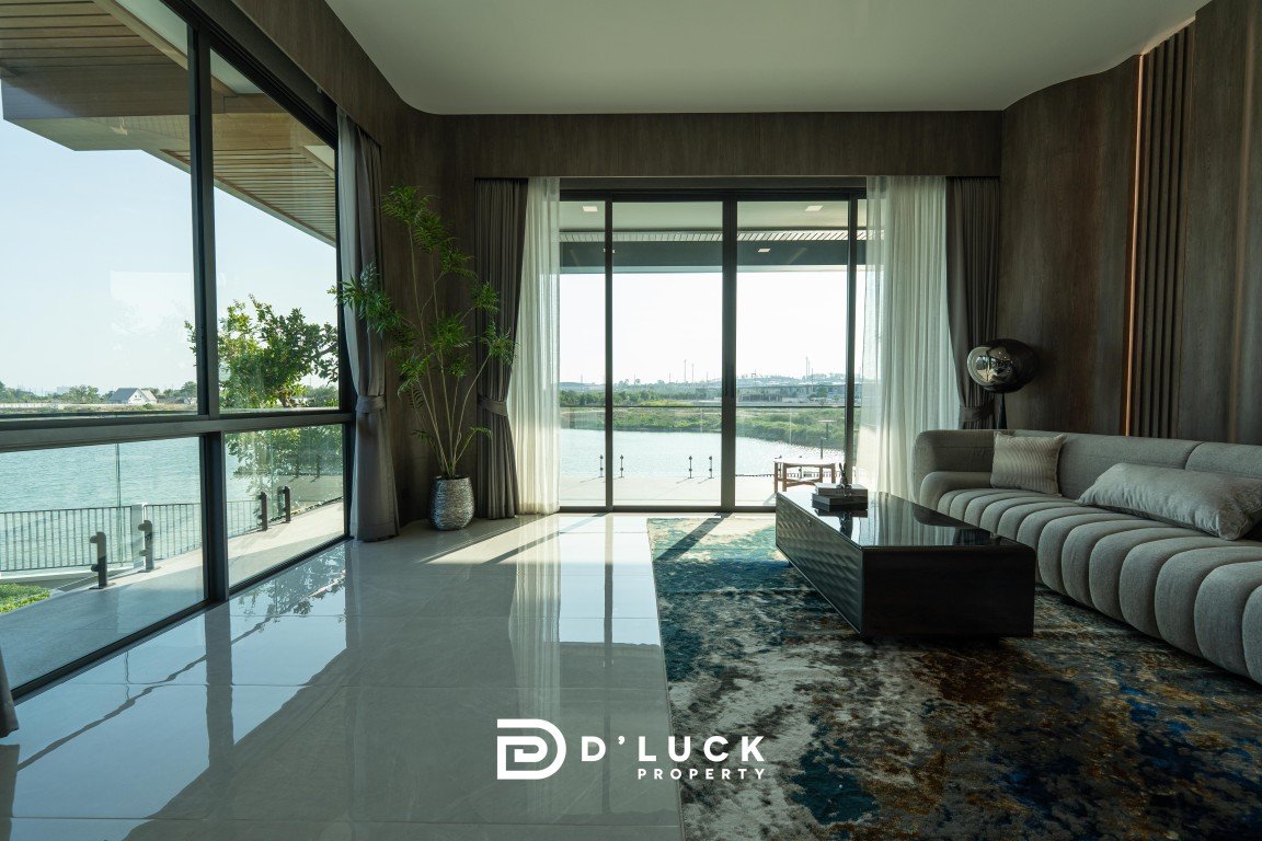 Bright modern living room with lake view through large windows, gray sectional sofa, glass coffee table, and a blue rug; D’Luck Property logo.