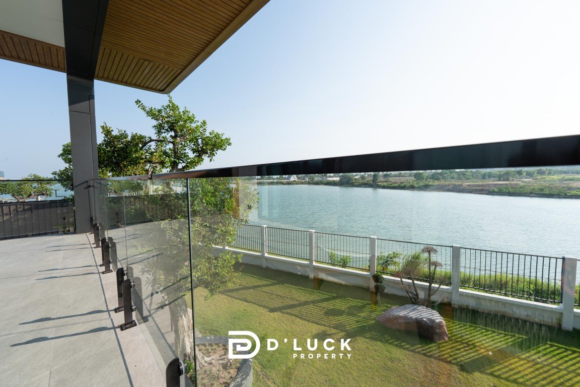 Modern balcony with glass railings overlooking a calm river, green lawn, and trees; D'LUCK Property logo at the bottom centre.