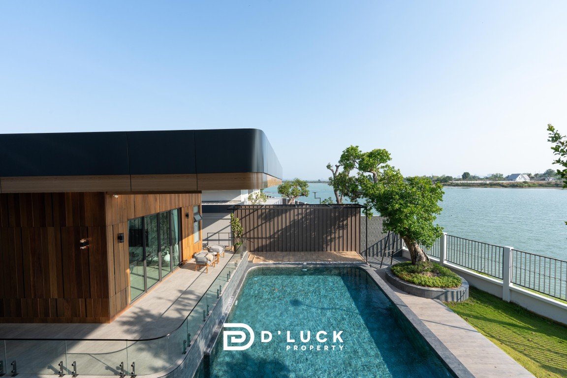 Modern contemporary house with wood cladding, glass railing, and a rectangular pool beside a waterfront view; includes 'D’Luck Property' logo in foreground.