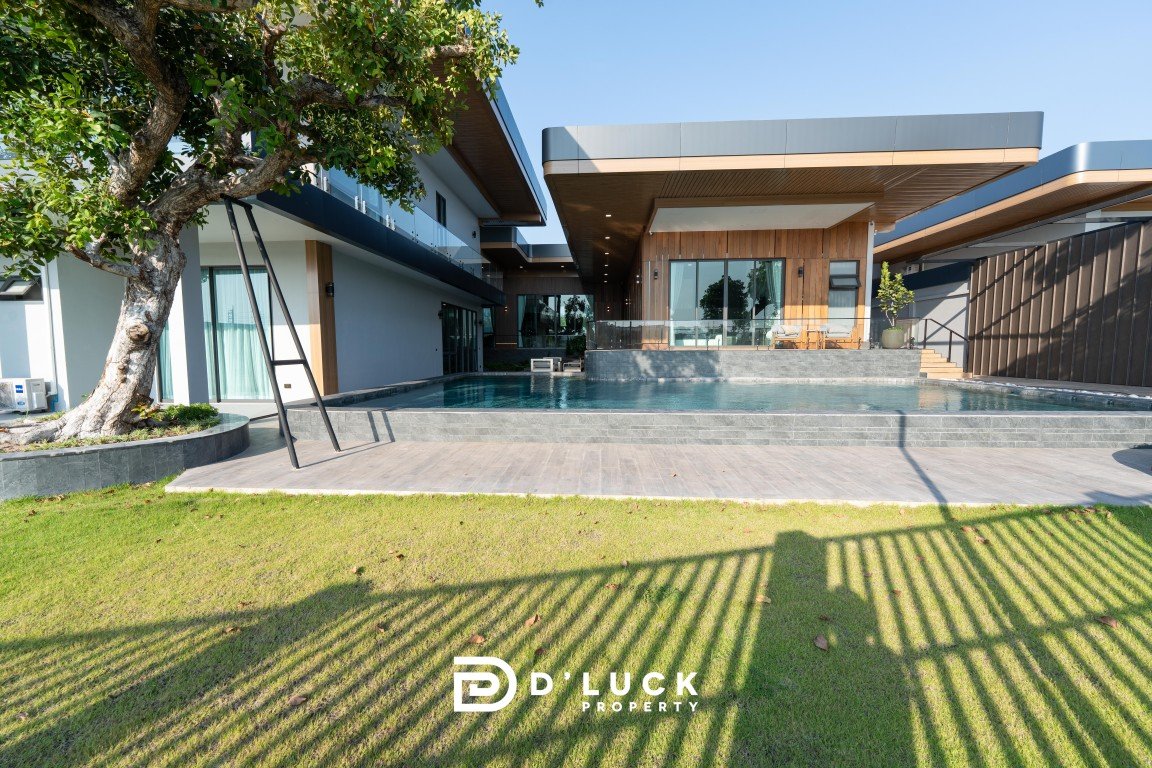 Contemporary villa with a rectangular pool, wooden facade, glass doors, and a D' Luck Property logo on the lawn.