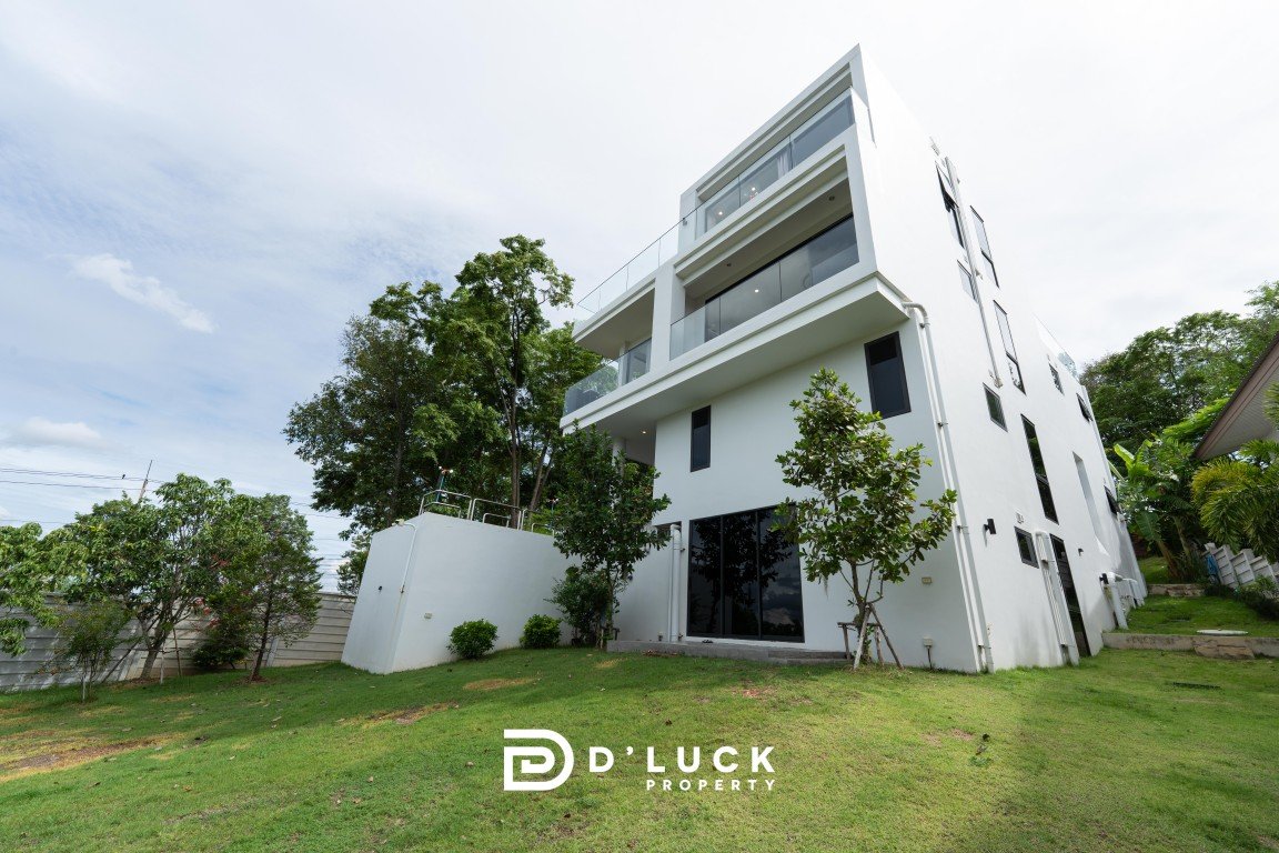 Modern white multi‑story house with glass balconies, set on a green lawn and surrounded by trees, under a blue sky.