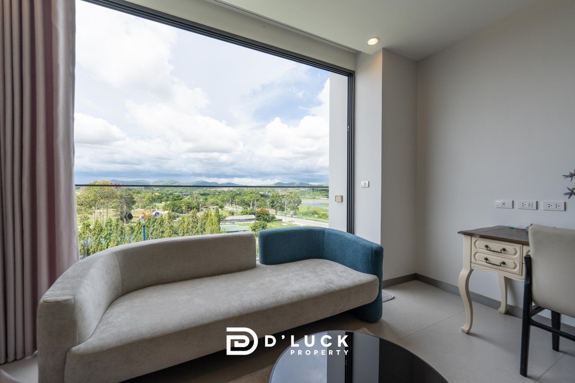 Modern living room with a beige curved sofa facing a large window showing a green landscape and distant hills.