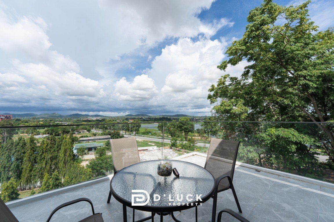 Balcony scene overlooking a valley and lake, with glass railing, a glass-topped table and three chairs, and a large tree on the right; watermark reads D'LUCK PROPERTY.