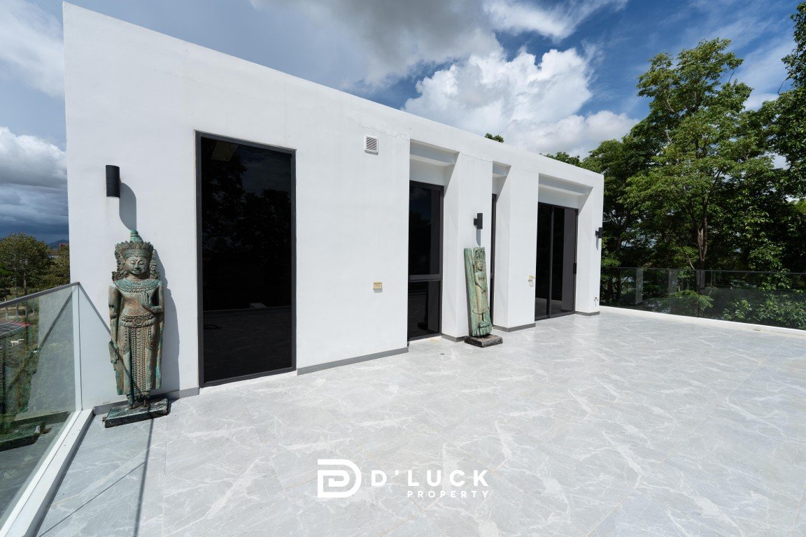 Modern white building with large glass doors on a marble terrace, flanked by two statue sculptures and glass railing; sunny day with trees in the background.
