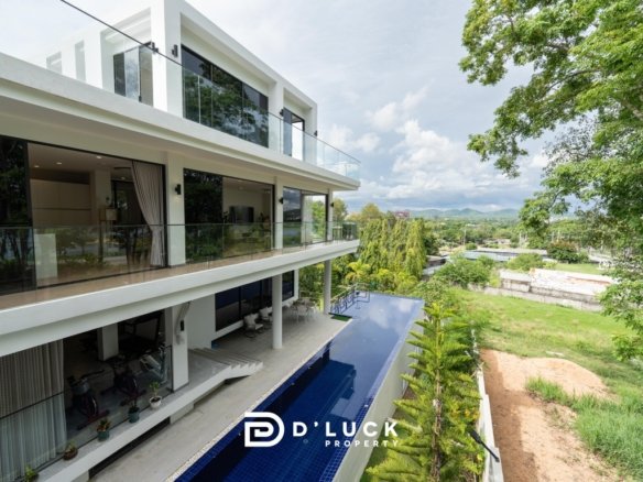 Modern white villa with glass balconies and a long blue pool set among green trees and hills in the distance with a sunny sky