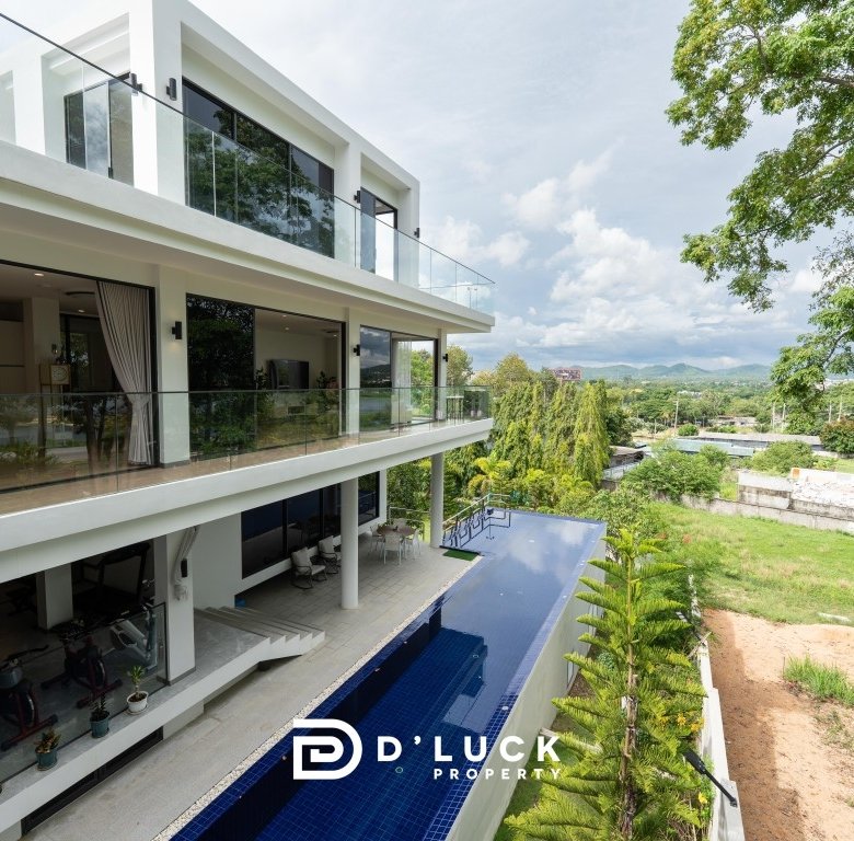 Modern white villa with glass balconies and a long blue pool set among green trees and hills in the distance with a sunny sky