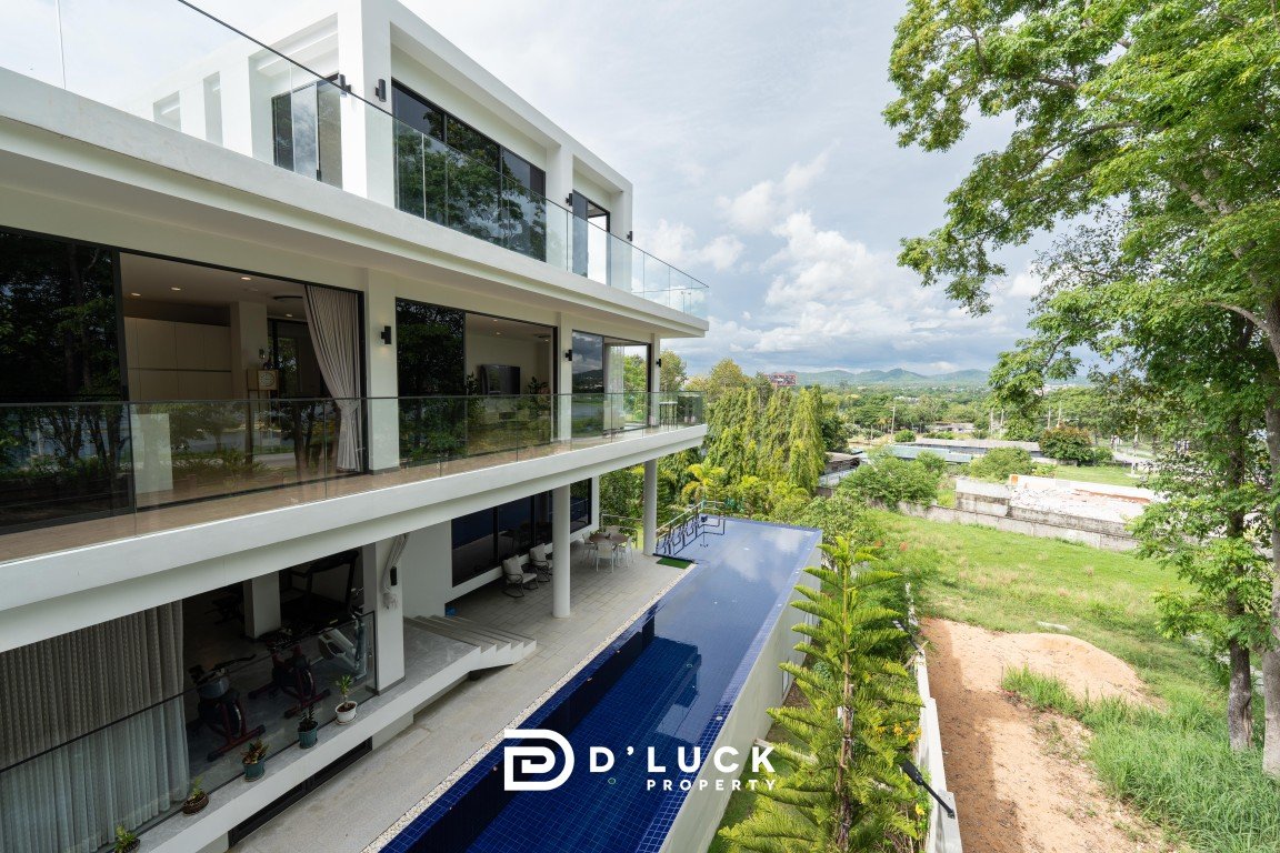Modern white villa with glass balconies and a long blue pool, set among green trees and hills in the distance, with a sunny sky.