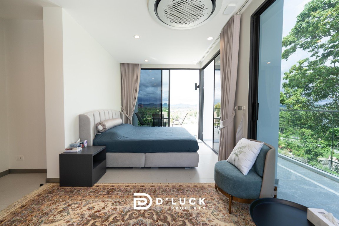 Bright hotel bedroom with a king bed, nightstand, and a seating chair, overlooking a balcony through large glass doors.