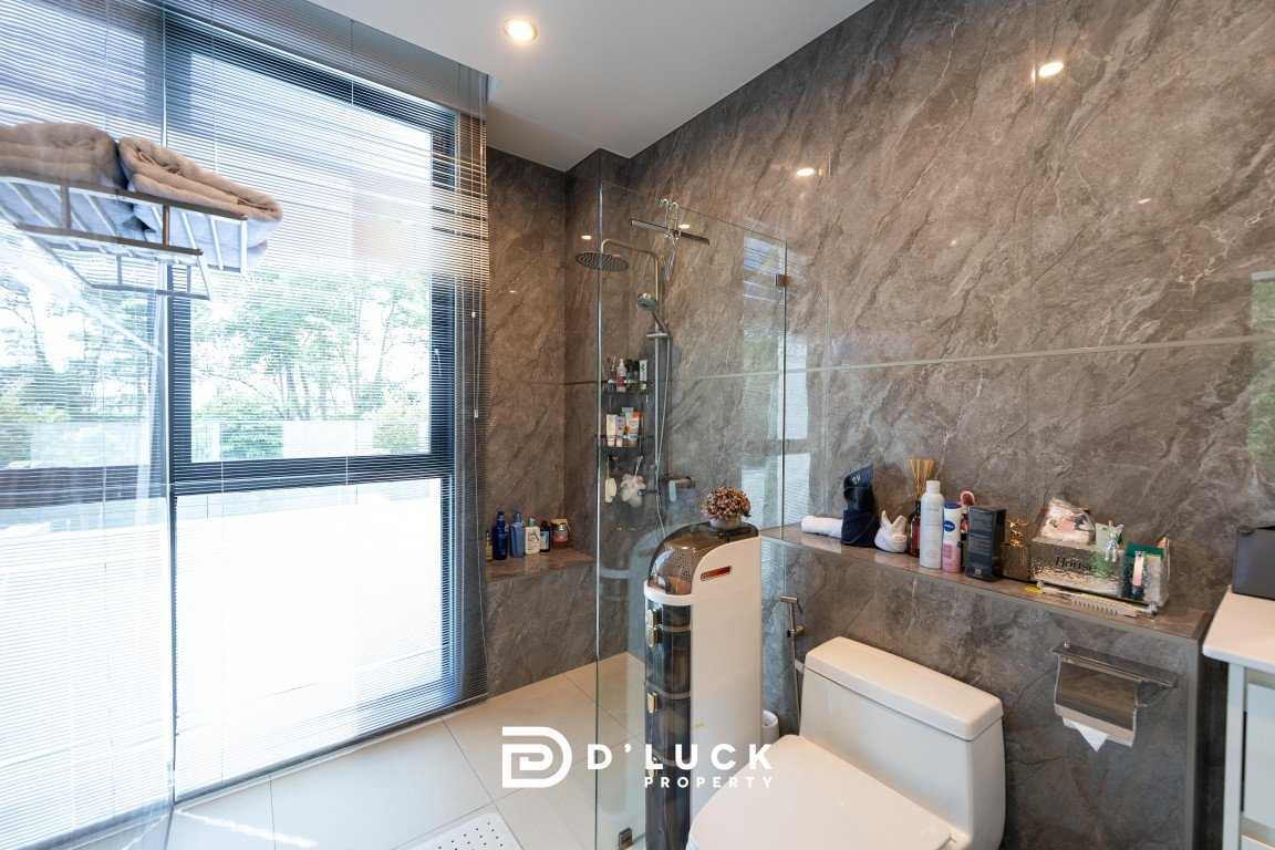 Modern bathroom with a glass shower enclosure, gray marble walls, and a toilet beside a ledge crowded with toiletries under a large window with blinds.