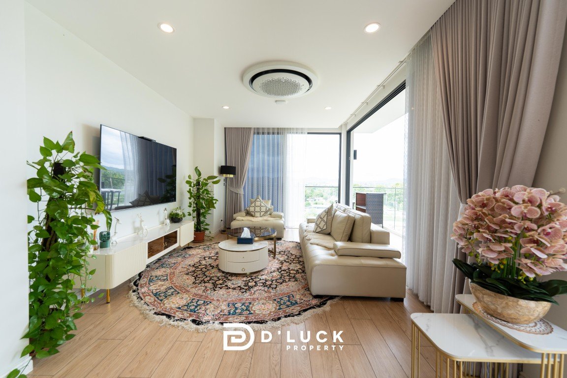 Bright living room with beige leather sofa, round coffee table, and a patterned rug; large windows with sheer curtains and multiple potted plants.