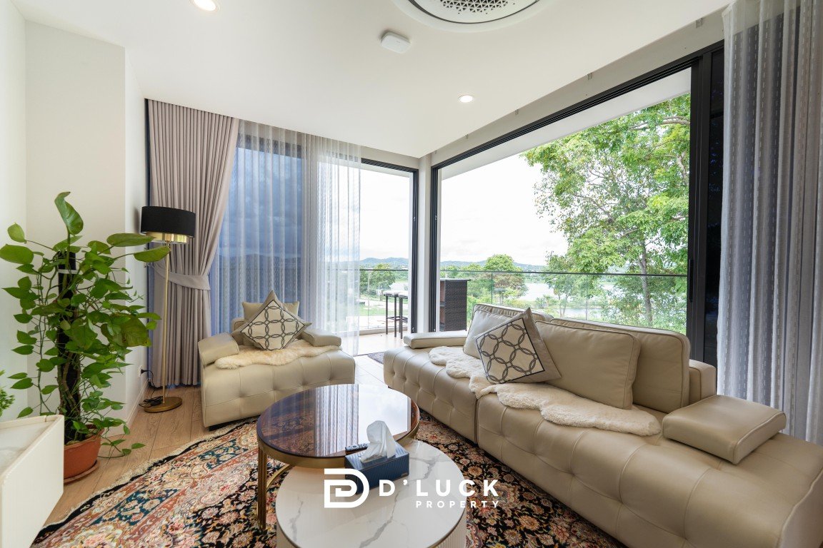 Bright living room with beige sectional sofas, a glass coffee table, and a patterned rug, opening to a balcony with green trees beyond (watermarked D'LUCK PROPERTY).