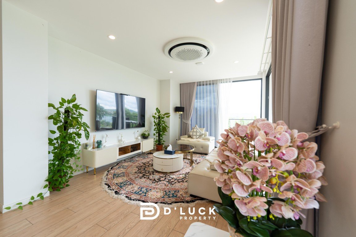 Bright living room with a wall-mounted tv, white media console, and green potted plants flanking a large rug and beige sectional sofa.