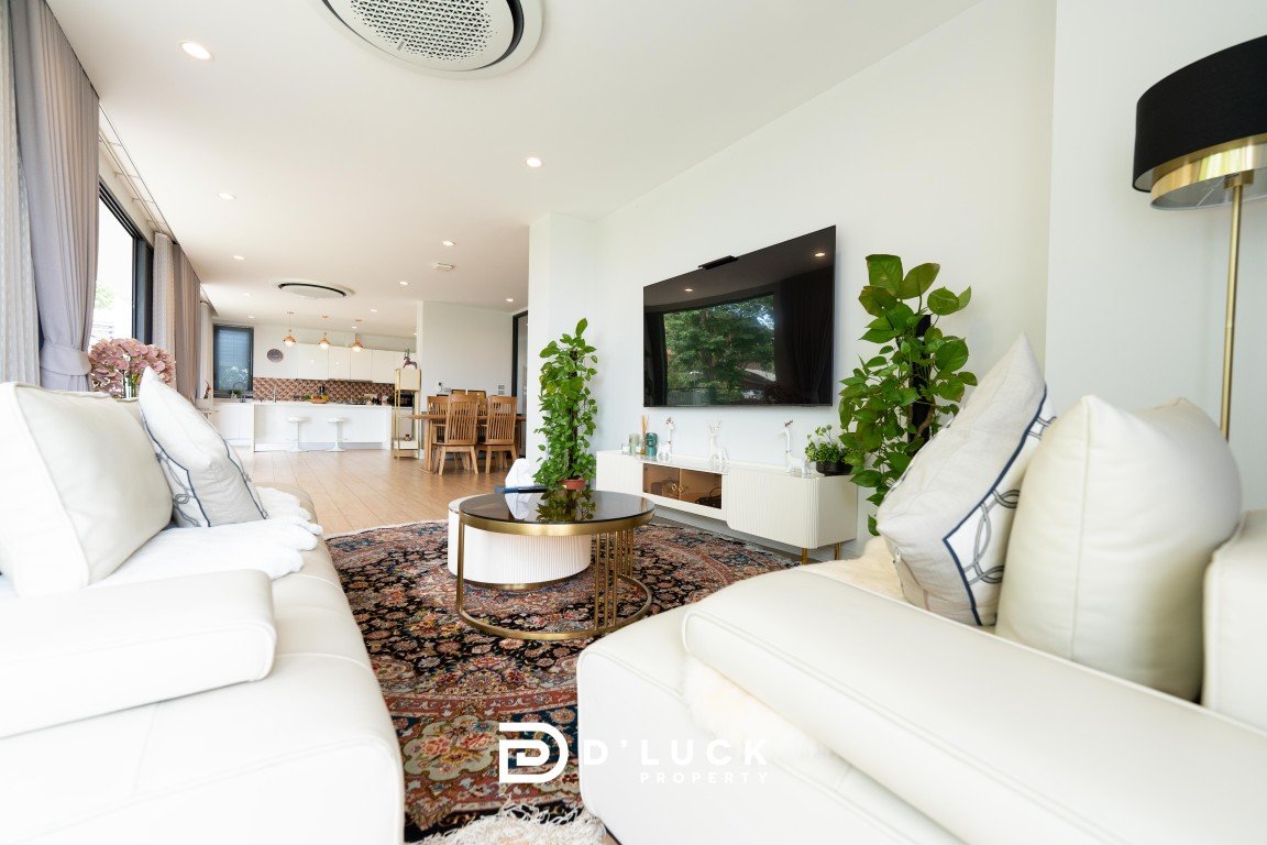 Bright open-plan living room with white sofas, glass coffee table, rug, wall-mounted TV, and potted plants; kitchen visible in the background.