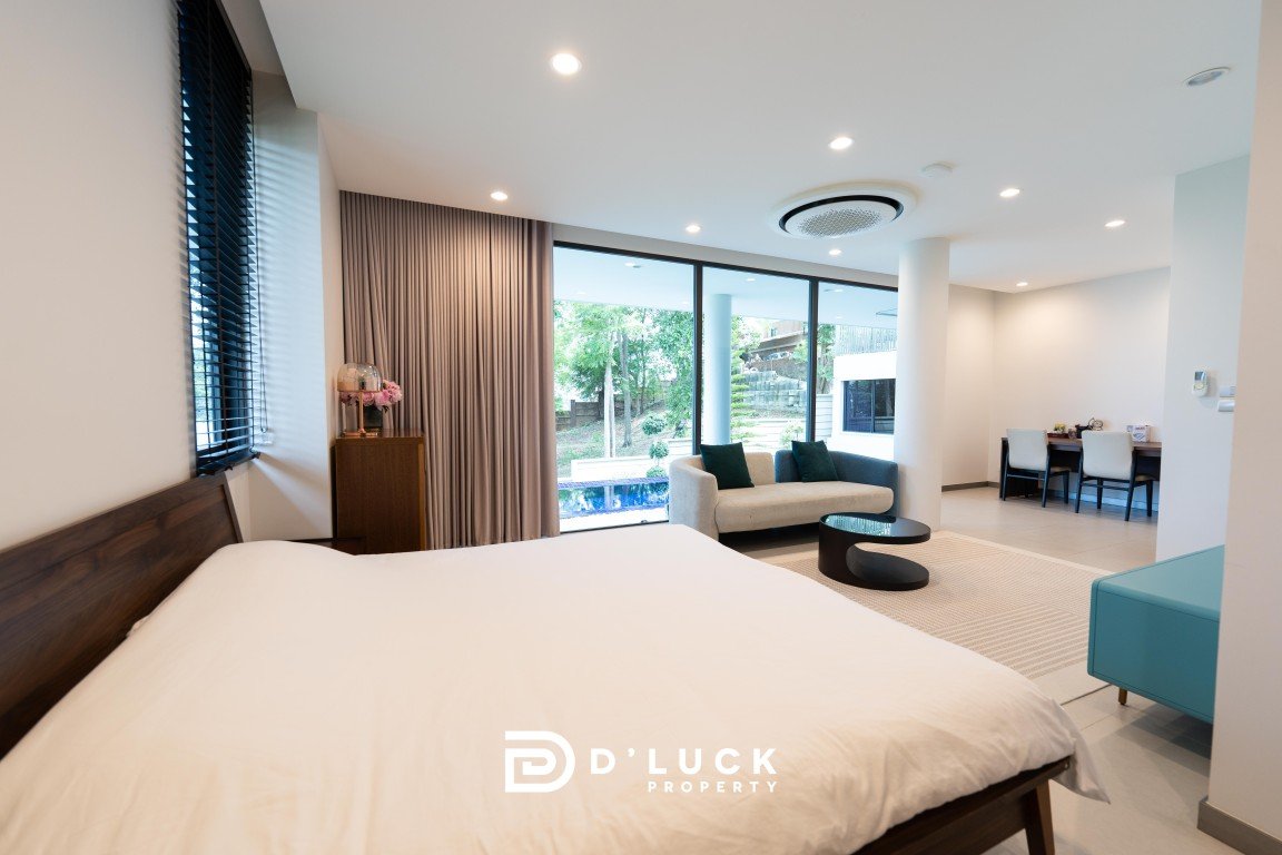 Modern hotel suite: large white bed in foreground, seating area by floor-to-ceiling windows, and glass doors to a green outdoor space; duvet bears the 'D’LUCK PROPERTY' logo.