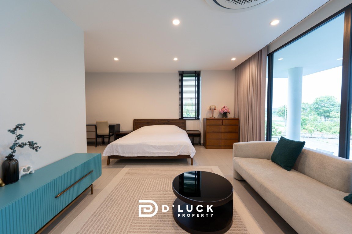 Modern hotel suite: centered bed, beige sofa, blue credenza, round black coffee table, and large windows with balcony view.