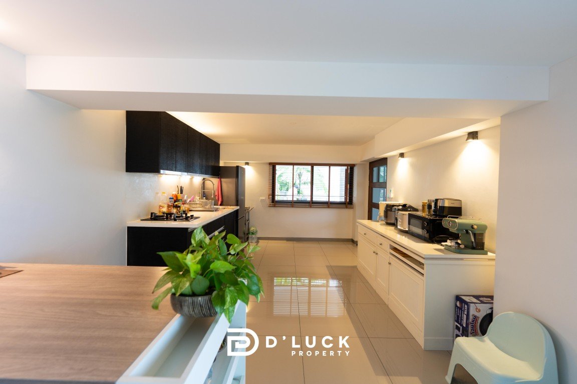 Bright open kitchen with white cabinets, black upper cabinets, and a row of appliances under a large window; potted plant in the foreground with D’LUCK PROPERTY logo.