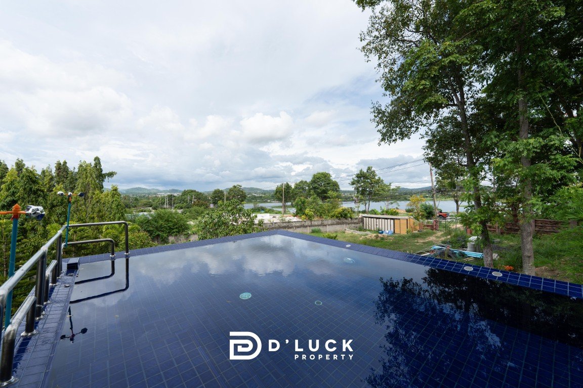 Infinity-edge pool reflecting the blue sky and clouds, surrounded by greenery and trees, with a distant landscape and a property logo at the bottom center.