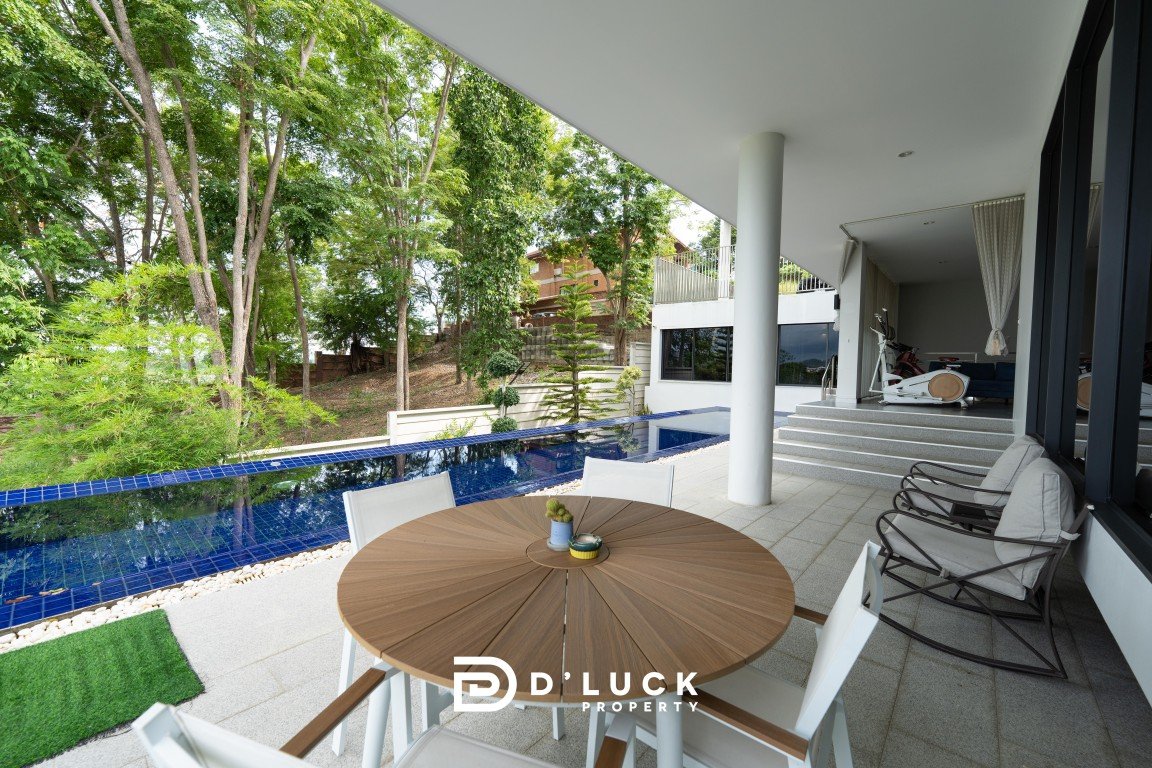 Outdoor patio with round wooden table and white chairs beside a narrow blue-tiled pool among trees.