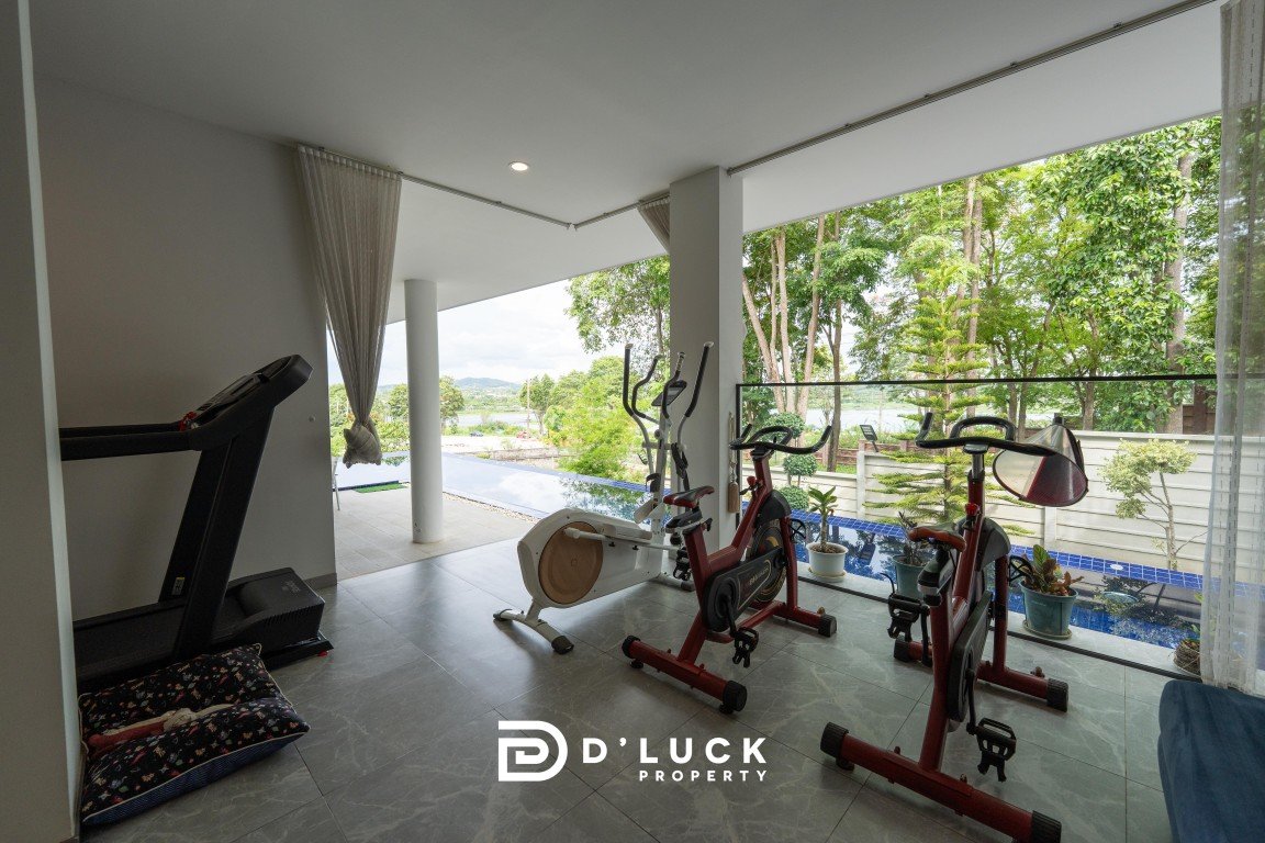 Modern home gym with a treadmill and two stationary bikes facing a covered patio and pool view beyond the glass railing, logo visible on floor.