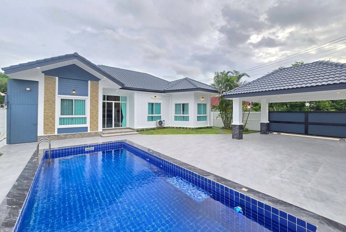 Modern white and blue house with a blue-tiled rectangular pool in the foreground under a cloudy sky, outdoor patio and glass doors visible.