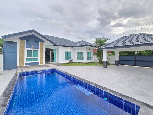 Modern white and blue house with a blue-tiled rectangular pool in the foreground under a cloudy sky, outdoor patio and glass doors visible.