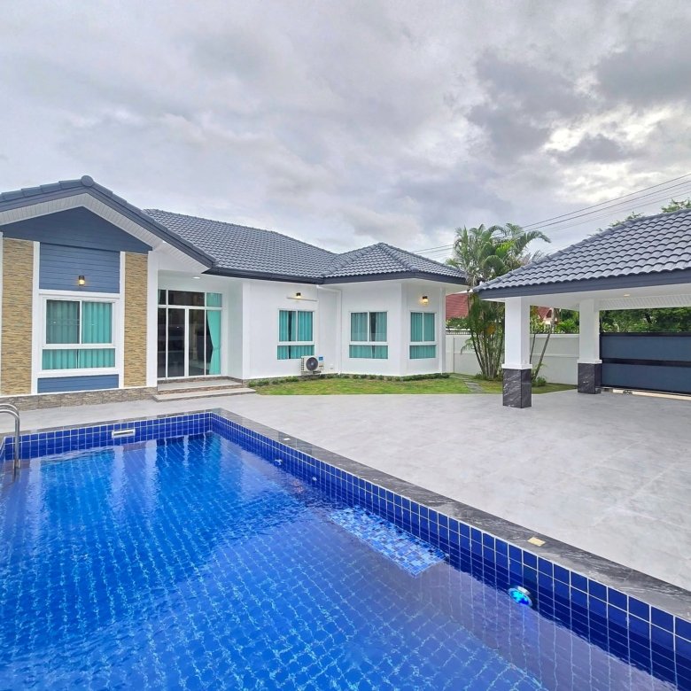 Modern white and blue house with a blue tiled rectangular pool in the foreground under a cloudy sky outdoor patio and glass doors visible