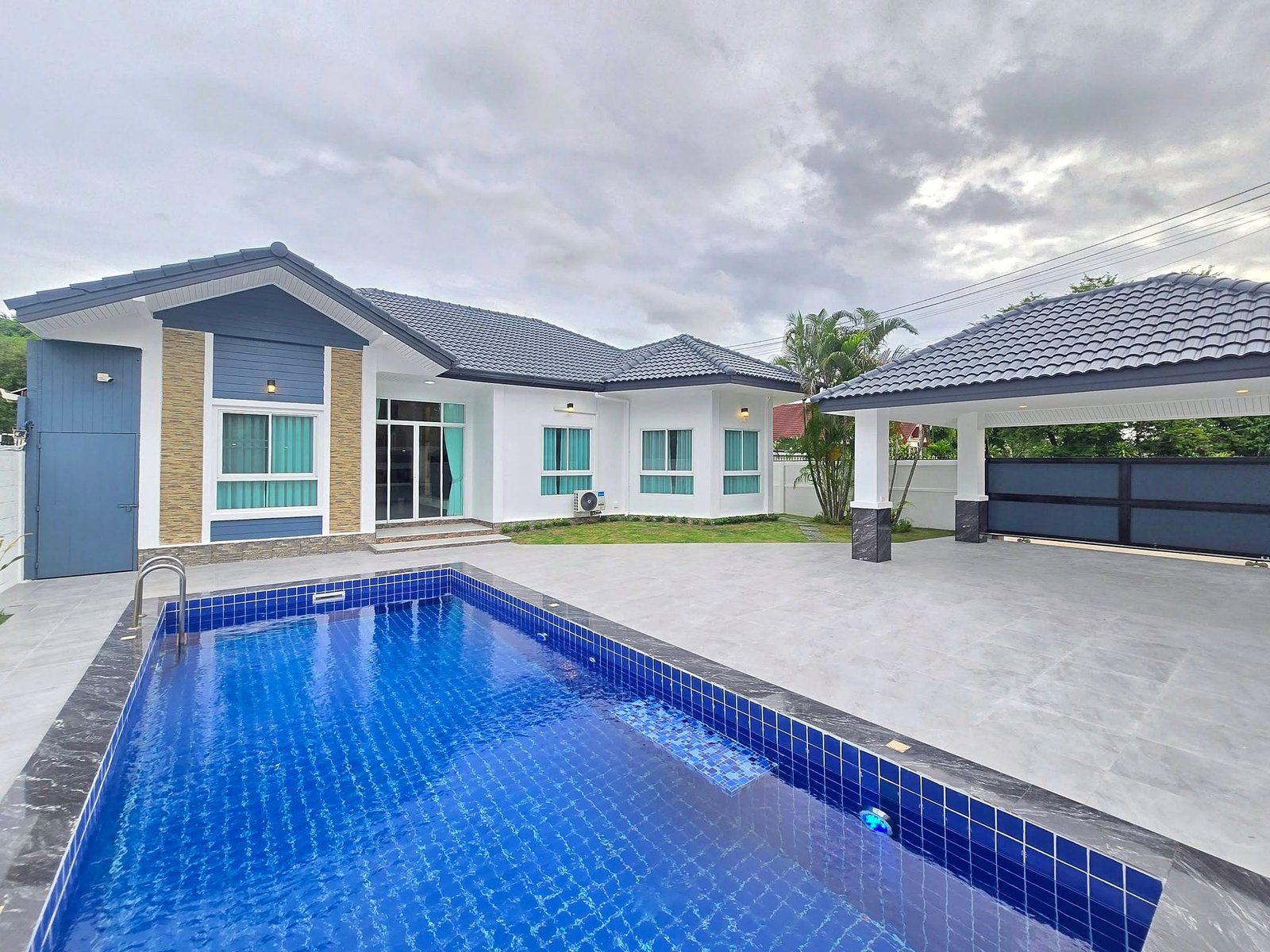 Mabprachan_Pool_Villa_Pattaya_Dlh571 Modern white and blue house with a blue-tiled rectangular pool in the foreground under a cloudy sky, outdoor patio and glass doors visible.