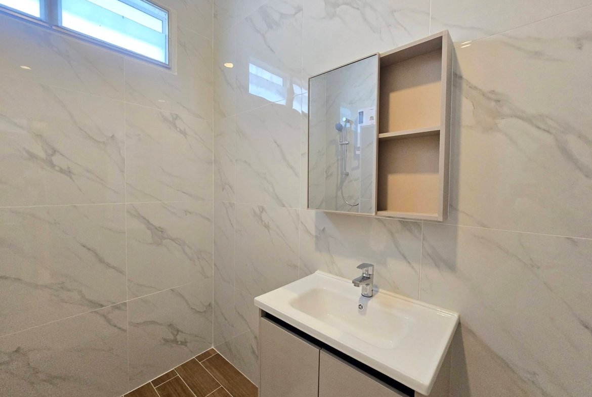 Modern bathroom with marble wall tiles, a white rectangular sink, and a mirrored vanity cabinet reflecting a shower area with a handheld showerhead. A high rectangular window adds natural light; wood-look floor tiles run along the corner.