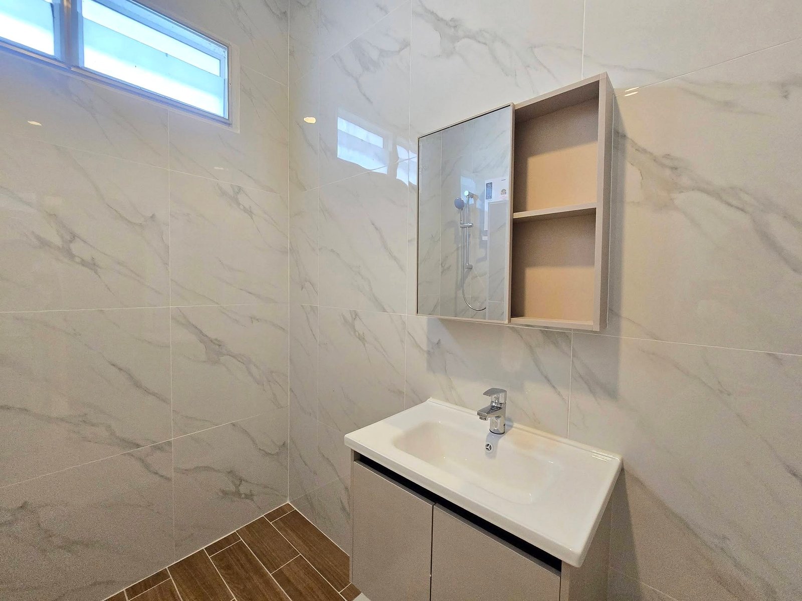 Mabprachan_Pool_Villa_Pattaya_Dlh571_11 Modern bathroom with marble wall tiles, a white rectangular sink, and a mirrored vanity cabinet reflecting a shower area with a handheld showerhead. A high rectangular window adds natural light; wood-look floor tiles run along the corner.