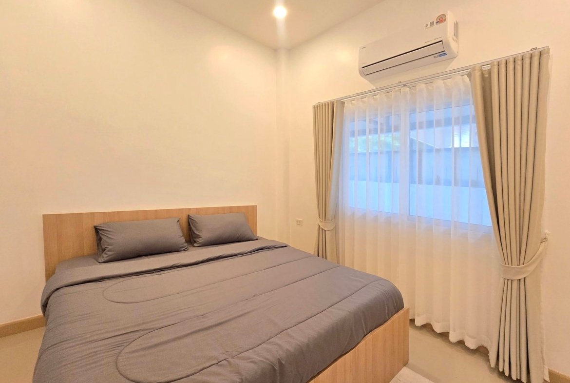 Bedroom with a wooden platform bed and gray bedding, sunlit window with sheer and beige curtains, and an air conditioner on the wall.
