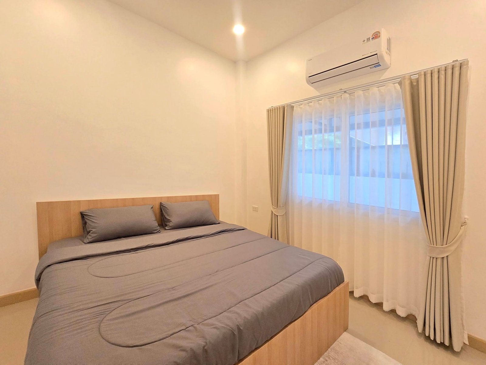 Mabprachan_Pool_Villa_Pattaya_Dlh571_12 Bedroom with a wooden platform bed and gray bedding, sunlit window with sheer and beige curtains, and an air conditioner on the wall.