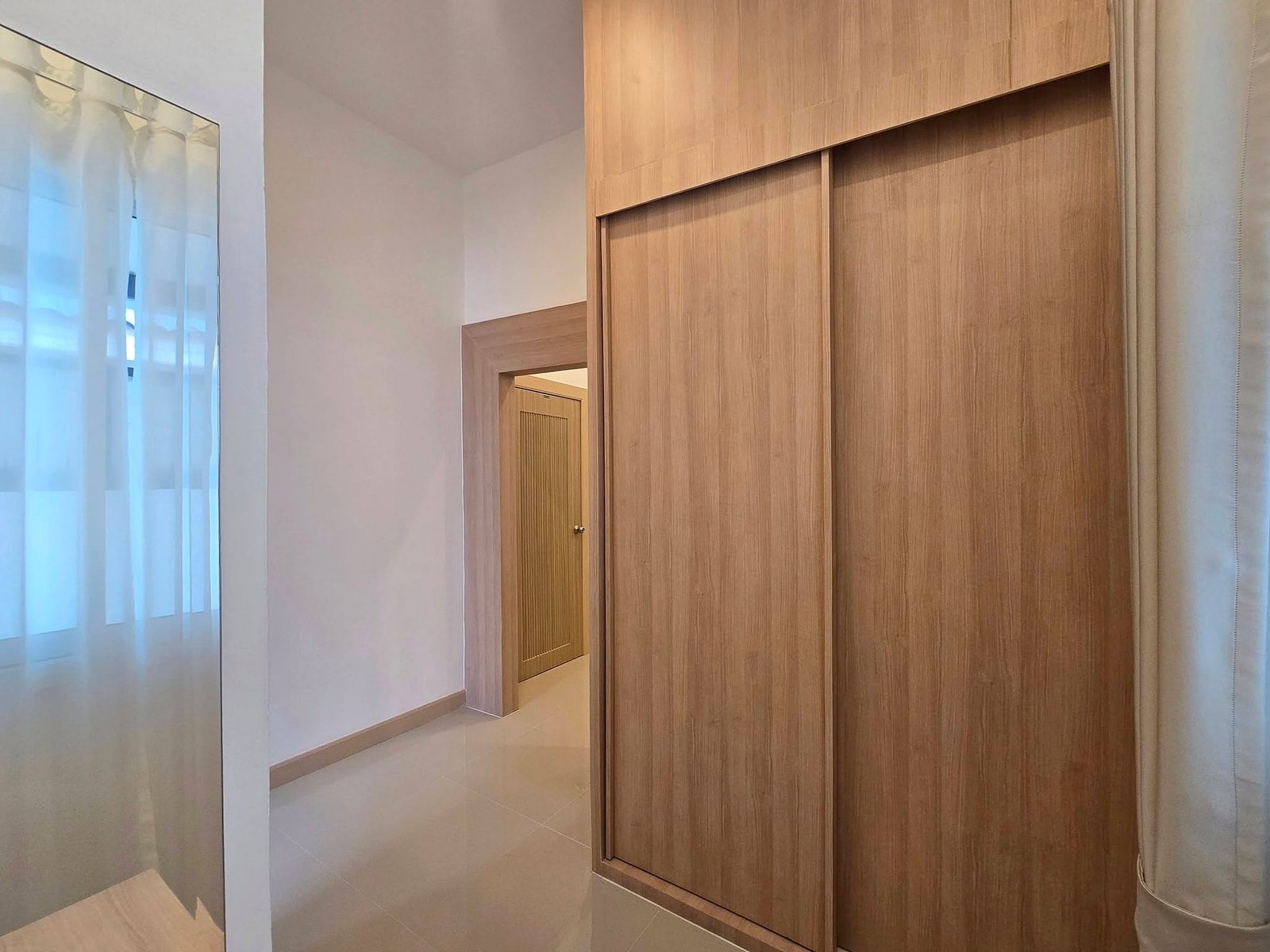 Mabprachan_Pool_Villa_Pattaya_Dlh571_13 Indoor hallway with a large wooden wardrobe featuring sliding doors on the right, doorway ahead, and sheer white curtains on the left.
