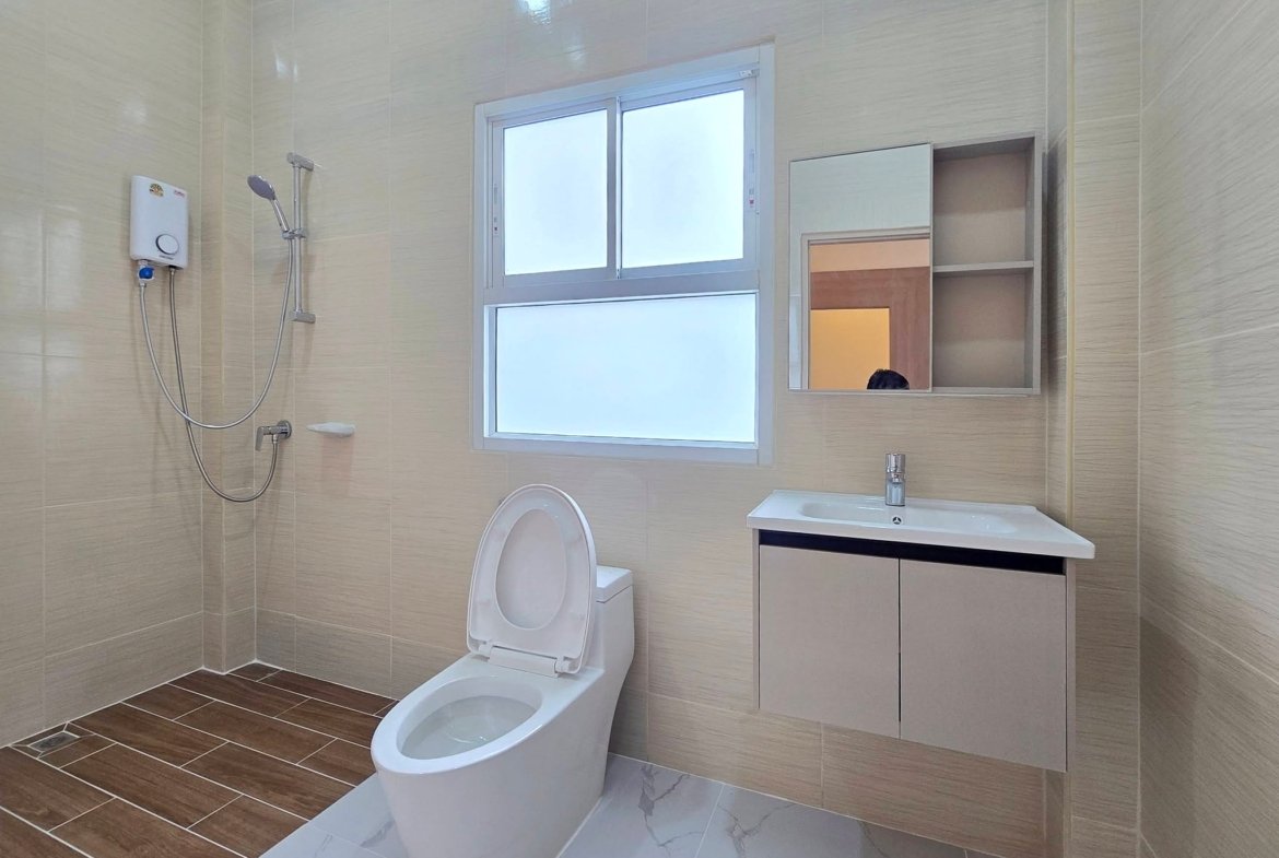 Modern bathroom with beige tiled walls, a wall-mounted handheld shower, white toilet, and a white vanity with sink beneath a window and mirror cabinet.
