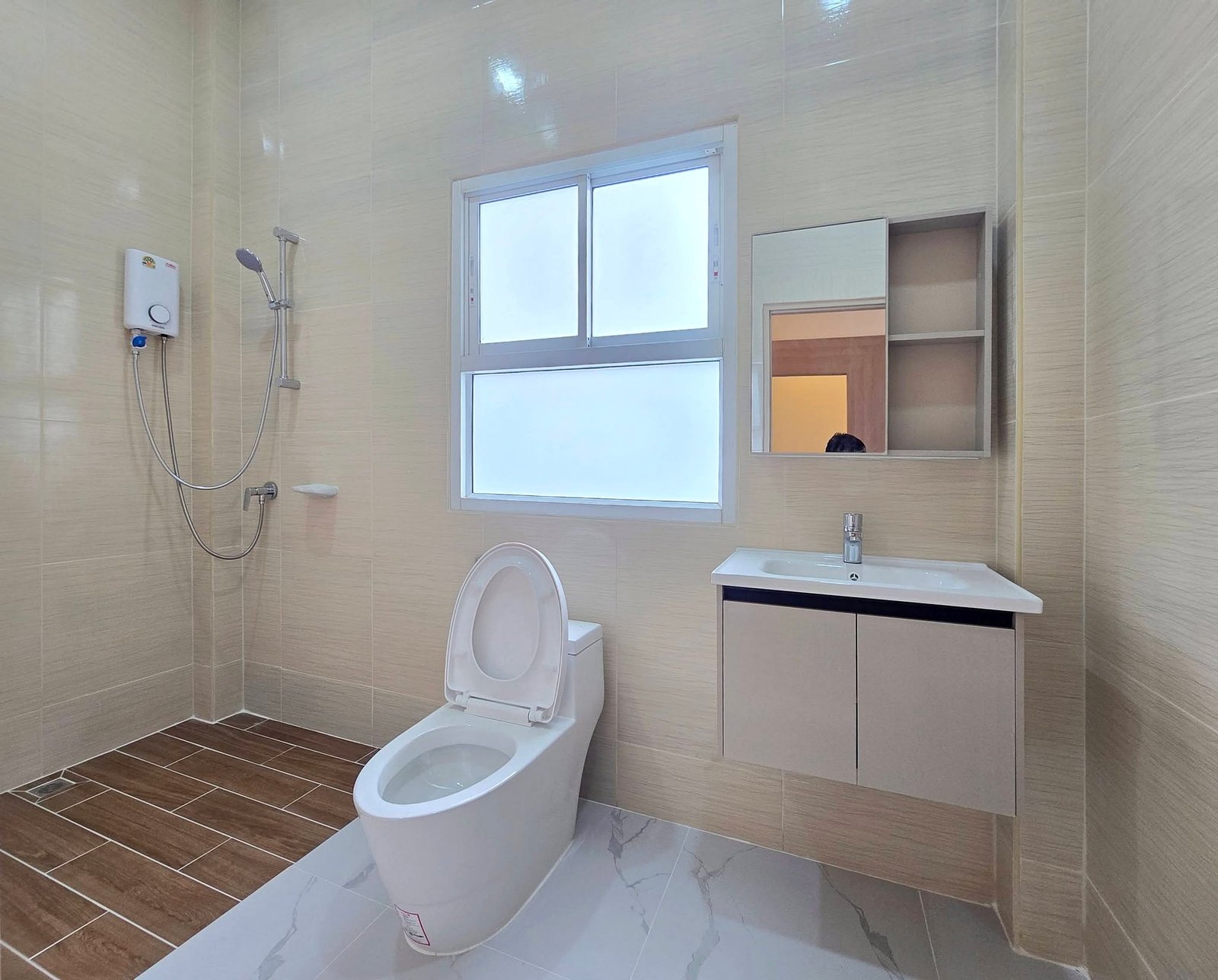 Mabprachan_Pool_Villa_Pattaya_Dlh571_14 Modern bathroom with beige tiled walls, a wall-mounted handheld shower, white toilet, and a white vanity with sink beneath a window and mirror cabinet.