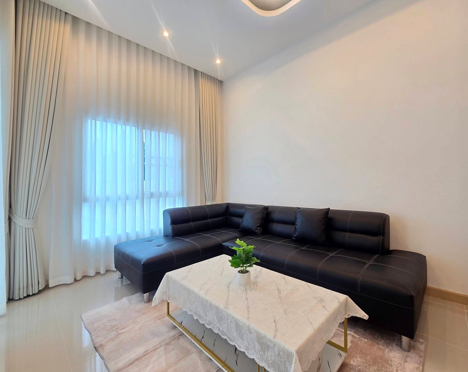 Mabprachan_Pool_Villa_Pattaya_Dlh571_15 Living room with a black leather sectional, white marble coffee tables with lace cloths, and tall windows with sheer curtains.