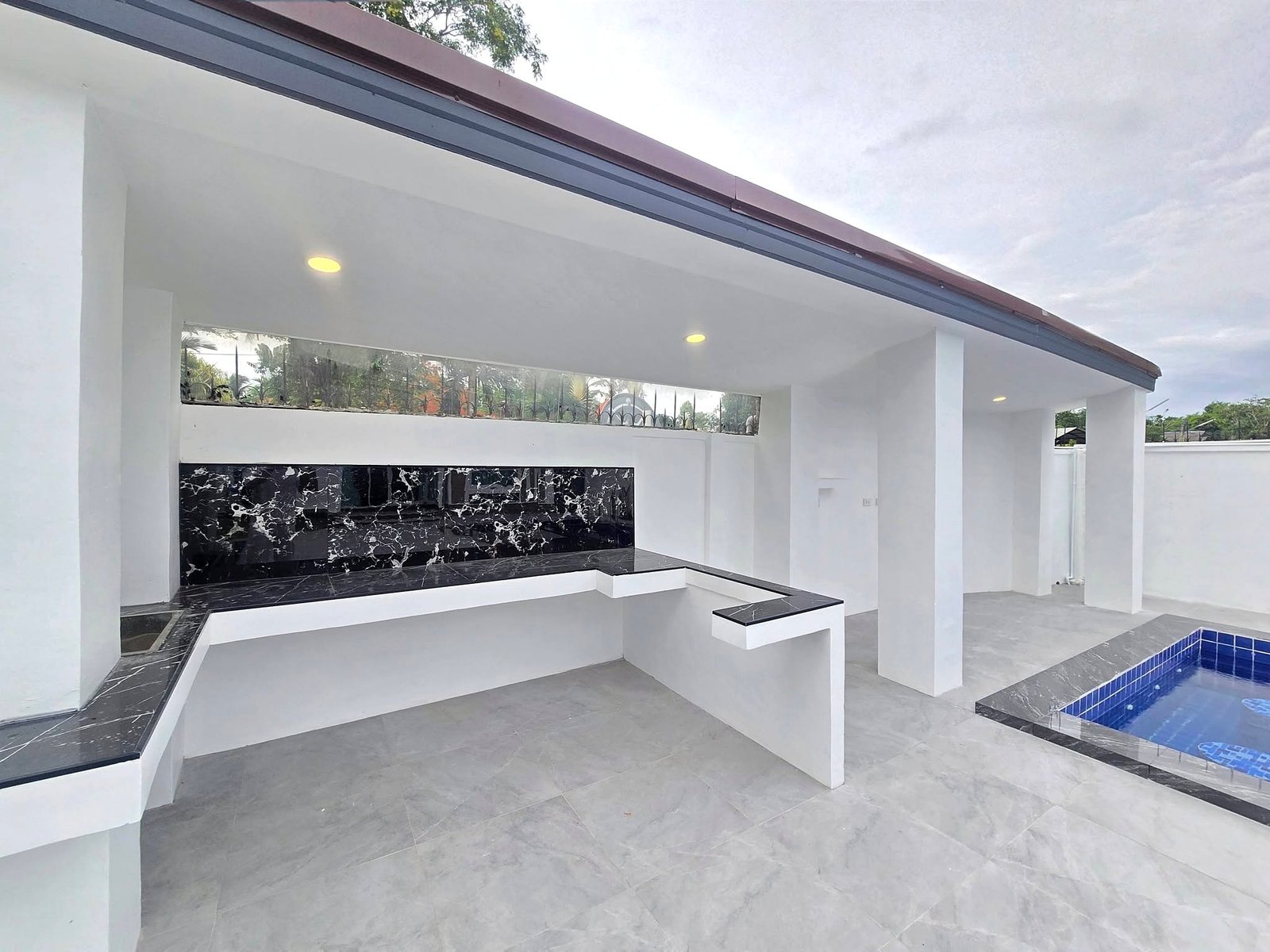 Mabprachan_Pool_Villa_Pattaya_Dlh571_17 Modern outdoor kitchen with white columns, black marble backsplash, and built-in countertop under a white roof; a blue-tiled pool is visible in the foreground/right.