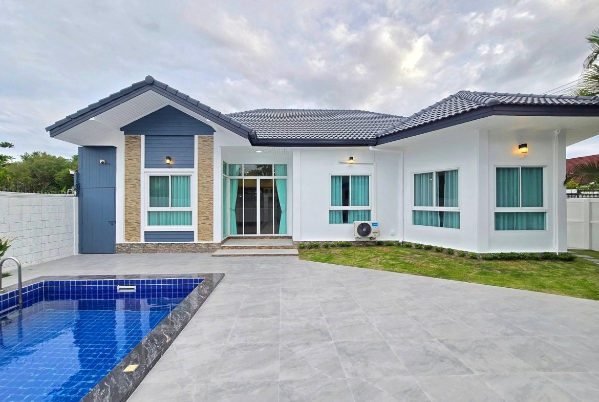Modern white house with blue accents and a blue-tiled pool in the foreground under a cloudy sky.